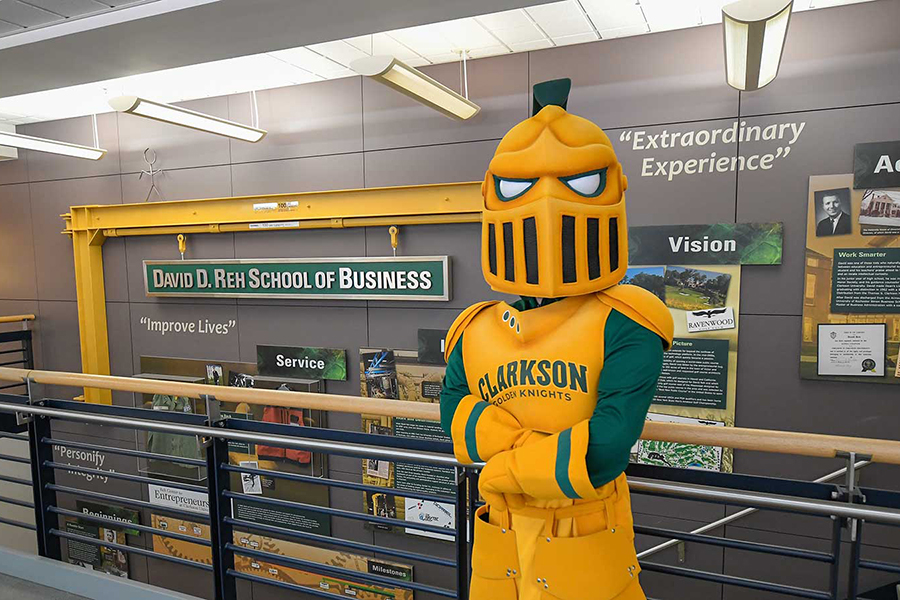 Clarkson's Golden Knight mascot posing with arms crossed in front of the Reh School of Business sign on campus